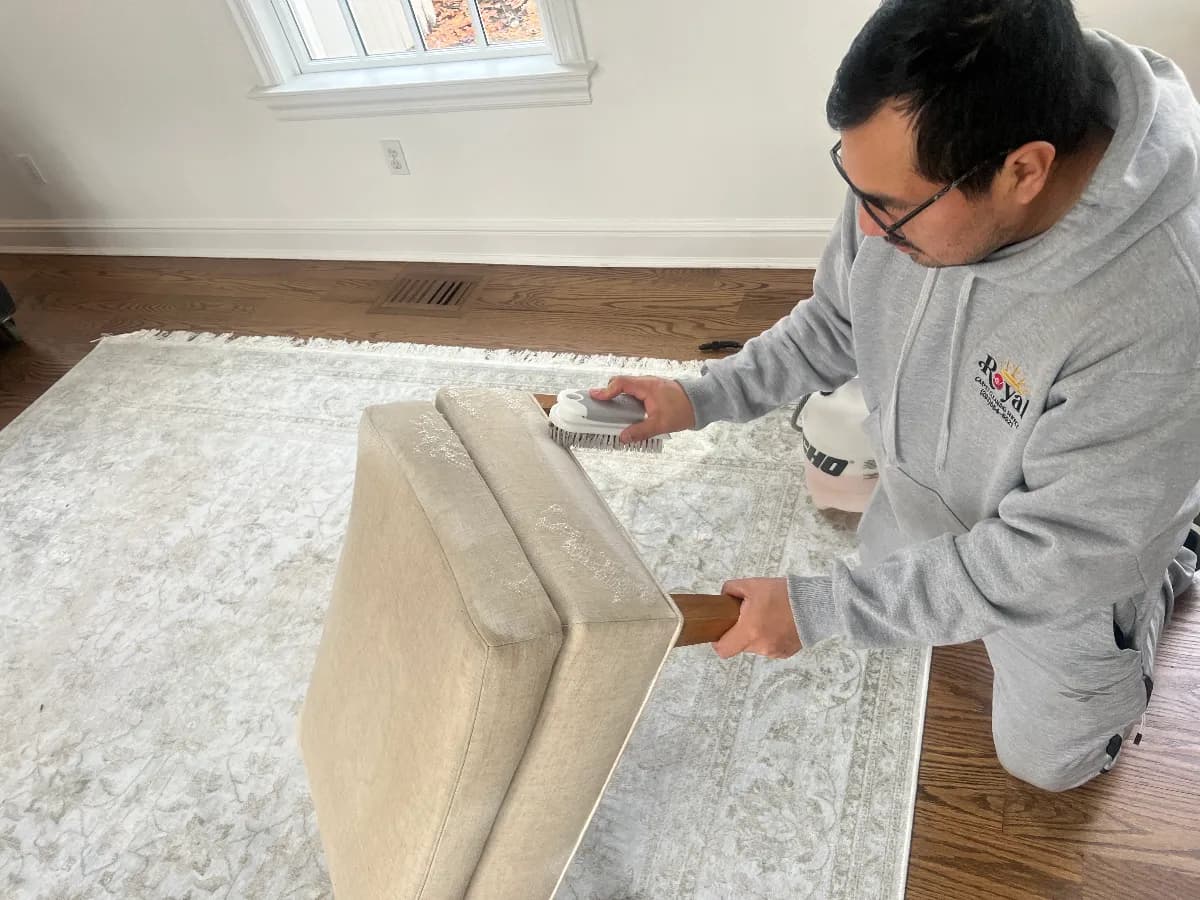 A Royal Carpet Cleaning technician meticulously scrubs a beige upholstered furniture piece using a professional handheld cleaning tool.